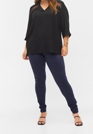 Top nero largo con scollatura a V e tessuto testurizzato, abbinato a pantaloni blu navy slim-fit e tacchi a strappy neri.