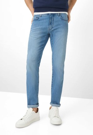 Homme portant un jean bleu clair retroussé, des baskets blanches et une chemise bleu marine, debout devant un fond blanc.