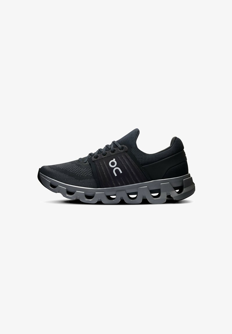 Baskets de sport noires avec un dessus en maille respirante, des accents sombres texturés et un design de semelle ondulée unique. Présente un logo sur le côté.