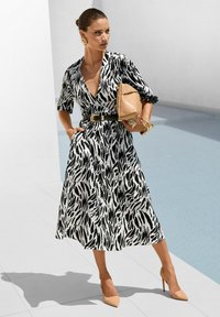 Schwarz-weiß gemustertes Wickelkleid mit kurzen Ärmeln, tailliert mit einem schwarzen Gürtel, kombiniert mit beigefarbenen spitzen Pumps und einer beige Handtasche.