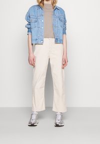 Chaqueta de mezclilla azul clara, camiseta beige, pantalones de pierna ancha color blanco roto y zapatillas plateadas. La chaqueta tiene bolsillos delanteros y botones.