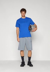 Blå kortermet t-skjorte med svart logo, grå sportsshorts, svarte joggesko og mørke sokker, holder en basketball med teksturert overflate.