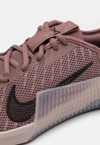 Zapatillas Nike en un suave tono malva con una parte superior de malla, logo Swoosh negro, paneles texturizados y una suela de goma translúcida. Presenta cordones planos y un collar acolchado.