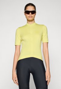 Kortermet sykkeltrøye i lys gul, med hel glidelås og tettsittende design, kombinert med svarte sykkelshorts.