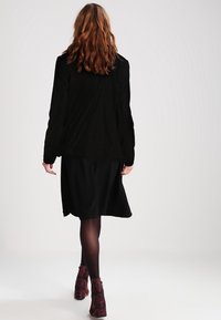 Černý sametový blazer s dlouhými rukávy a přizpůsobeným střihem, doplněný černou sukní po kolena, nošený s vzorovanými kotníkovými botami.