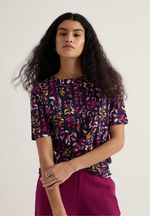 Woman with long curly hair wearing a navy blue floral short-sleeve top and dark pink pants, standing with arms crossed against a plain background.