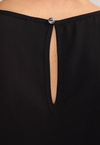 Haut noir avec une ouverture en forme de clé à l'arrière, fermée par un bouton métallique. Le tissu semble lisse et léger.