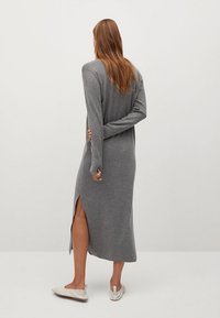 Femme aux cheveux longs portant une longue robe en tricot grise avec une fente sur le côté, debout pieds nus sur un fond blanc uni.