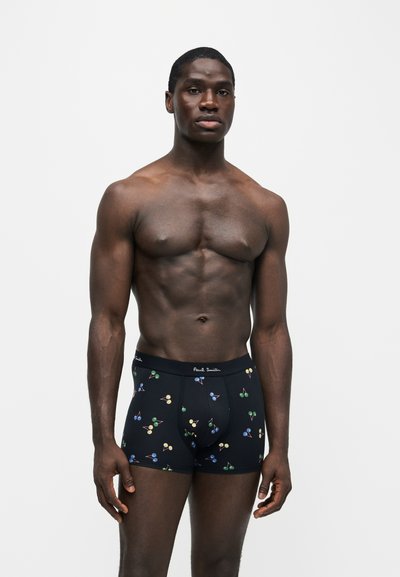 Modèle masculin fort à la peau foncée portant un boxer noir avec des motifs colorés de cerises sur fond blanc uni.