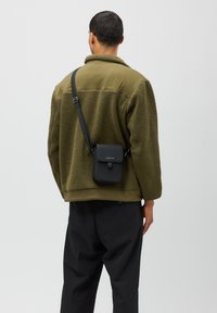 Svart läder crossbody-väska med klaffstängning, fäst vid ett svart band, mot en bakgrund av en grön fleecejacka och mörka byxor.