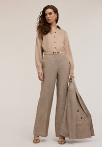 Beige långärmad skjorta med svarta knappar, i kombination med ljusbruna randiga byxor och en matchande kavaj. Modellen håller kavajen.