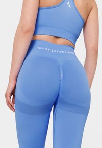 Modré fitness legíny majú vysoký pás, textúrované akcenty a branding na páse. Tkanina sa zdá byť hladká a priliehavá.