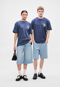 Tamsiai mėlynos per dideli marškinėliai su apskritu logotipu, derinami su šviesiai mėlynais džinsiniais šortais. Juodos spalvos aksesuarai ir batai užbaigia aprangas.