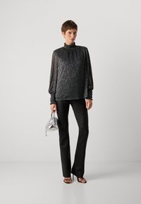 Top lungo nero texturizzato con colletto alto a volant, abbinato a pantaloni neri slim e una piccola borsa a tracolla argentata.