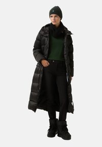 Doudoune noire, silhouette longue, écharpe noire côtelée, pull vert, pantalon noir, bottes d'hiver noires et bonnet vert avec détail de logo.