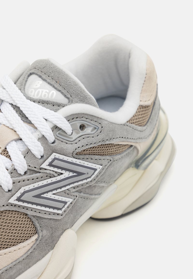 Baskets grises avec une tige en mesh et en daim, lacets blancs, et une semelle à amorti visible. Présente un logo 'N' bien en évidence et une marque sur la languette.