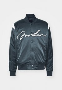 Svart sateng bomberjakke med hvite detaljer og stor "Jordan" signaturlogo på brystet. Har trykknap-lukking og ribbestrikkede ermer.
