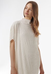 Witte textuur jurk met ingewikkelde reliëf, met een hoge halslijn en korte mouwen, die cirkelvormige en lineaire patronen toont.