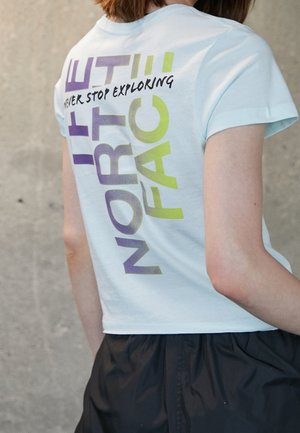 Osoba nosząca biały krótki T-shirt z tekstem "THE NORTH FACE" i "NEVER STOP EXPLORING" w kolorze fioletowym i zielonym na plecach.