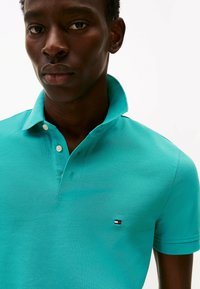 Türkises Polo-Shirt aus strukturiertem Stoff, mit klassischem Kragen, zwei Knöpfen und einem kleinen Logo-Patch in Rot, Weiß und Blau.