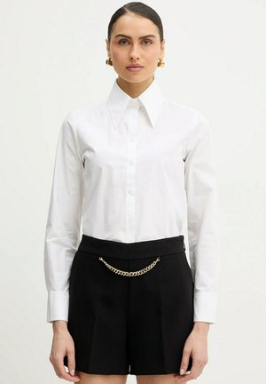 Donna che indossa una camicia bianca con bottoni, pantaloncini neri a vita alta con dettaglio di catena dorata e orecchini a cerchio dorati, in piedi contro uno sfondo neutro.