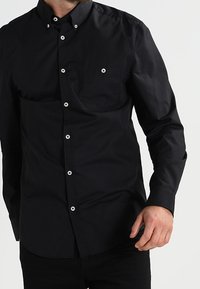 Camicia nera con colletto, maniche lunghe e taschino. Presenta bottoni bianchi e una texture di cotone liscia.
