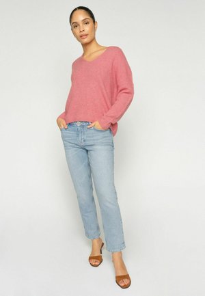 Woman standing with hands in light blue jeans pockets wearing a pink V-neck sweater and brown open-toe heeled sandals against a white background.