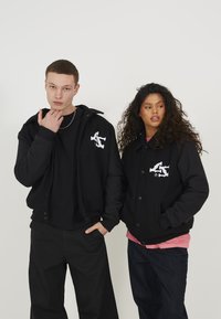 Zwarte fleece jassen met knoopsluitingen, voorzien van witte grafische ontwerpen. Modellen dragen losse zwarte broeken en gelaagde tops.