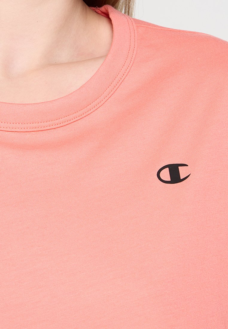 T-shirt à manches courtes de couleur pêche, au design col rond, fabriqué en tissu doux. Présente un petit logo noir sur la zone de la poitrine gauche.