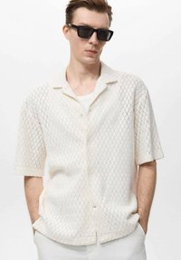 Camisa blanca de manga corta con un diseño texturizado de punto abierto. Presenta un cuello tipo polo y cierre de botones, utilizada sobre una capa ligera.