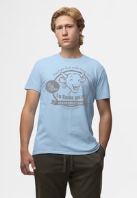 Světle modré bavlněné tričko s grafikou usmívající se krávy a šedým textem, umístěné na neutrálním pozadí.