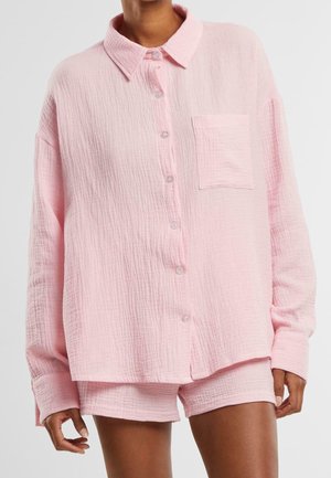Person iført løs tekstureret lyserød skjorte med brystlomme og matchende shorts mod en ensfarvet baggrund.