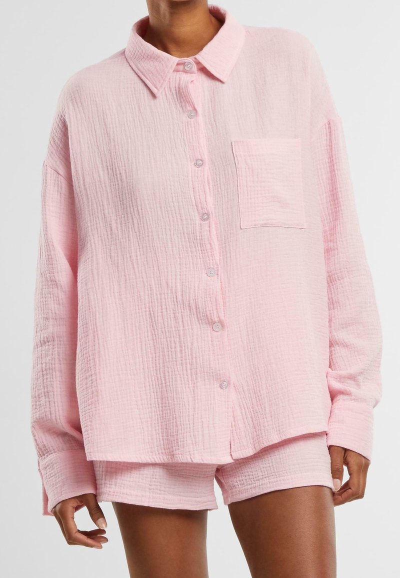 Personne portant une chemise rose pâle texturée, ample, boutonnée avec poche poitrine et un short assorti sur un fond uni.