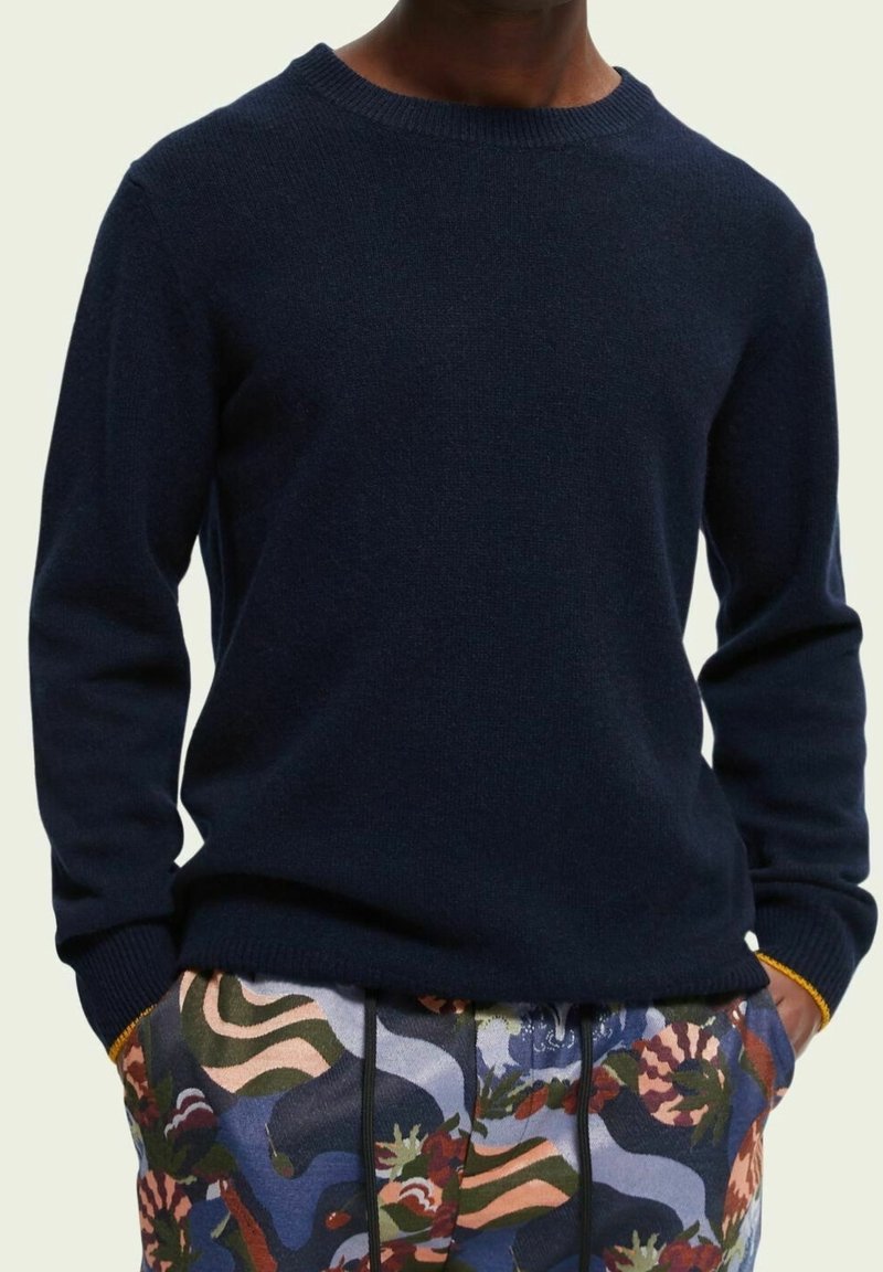 Navyblauwe gebreide sweater met een ronde hals en ribbels aan de mouwen, gecombineerd met gemusterde, multicolor broek met bloem- en golfpatronen.