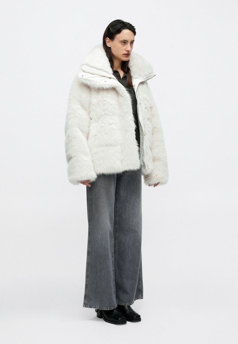 Doudoune en fausse fourrure blanche avec col montant, fermeture éclair à l'avant et coupe oversize, associée à un jean gris à jambes larges et des bottes noires.