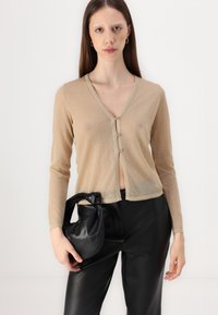 Beige, gennemsigtig cardigan med lange ærmer og V-hals, der har tre knapper foran og en struktureret overflade. Båret med sorte læderbukser og taske.