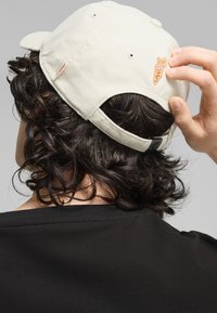 Beżowa czapka z wygięnym daszkiem, regulowanym paskiem i pomarańczowym haftem marchewki z tyłu; noszona na kręconych ciemnych włosach i czarnej koszulce.