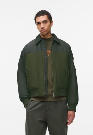 Green bomber jacket with a zip closure, ribbed cuffs, and a two-tone design featuring a textured upper section and smooth lower section.
