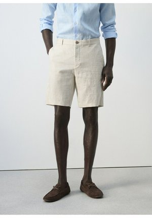 Uomo con pantaloncini di lino beige, camicia azzurro chiaro con maniche arrotolate e mocassini in camoscio marrone scuro in piedi su un pavimento grigio.