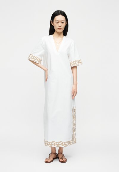 Femme portant une robe blanche jusqu'aux chevilles avec des motifs brodés beiges sur les manches et l'ourlet, associée à des sandales plates marron.