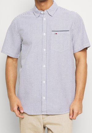 Man wearing short-sleeve, light blue and white vertical striped button-up shirt with a small front chest pocket and beige pants.