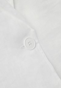 Szczegół białej lnianej koszuli z białym plastikowym guzikiem na cztery otwory i delikatnym przeszyciem, z gładką, teksturowaną fakturą.