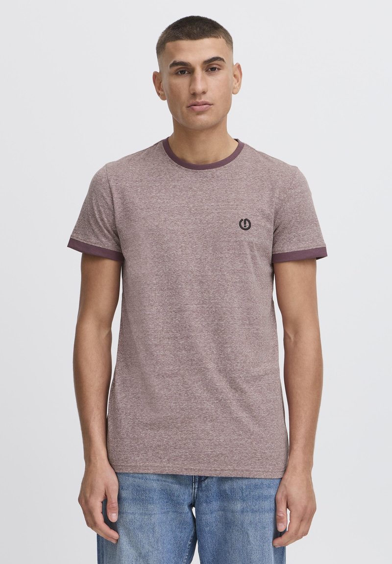 Camiseta de manga corta color marrón con un cuello y remates de manga en un marrón oscuro en contraste; presenta un pequeño logo negro en el pecho.