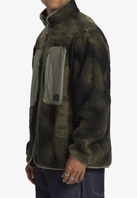 Chaqueta de forro polar en verde oliva con un acabado texturizado, que presenta un bolsillo en el pecho con cremallera y acentos de tela lisa en contraste.