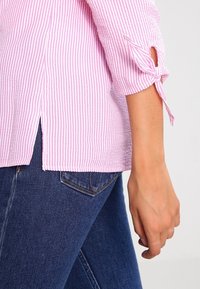 Světle růžová a bílá pruhovaná košile s tříčtvrtečními rukávy, s uzlem na rukávu a rozparkem na boku.