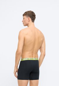 Čierne boxerky s vzorovaným šedým a neonovo zeleným pásom v prednej časti s logom "NIKE", zobrazené zozadu. Hladký povrch.