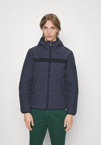 Giacca a vento navy trapuntata con cappuccio e chiusura con zip, caratterizzata da una striscia nera orizzontale e dal marchio "HILFIGER". Tessuto leggero, tasche laterali.