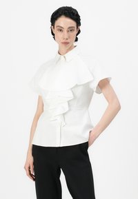 White blouse featuring a ruffled front design, short sleeves, and a structured collar. Smooth fabric with subtle sheen. Paired with black pants.