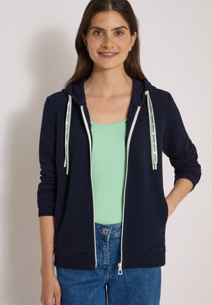 Femme souriante portant un sweat à capuche bleu marine zippé avec des cordons blancs, un haut vert clair et un jean bleu, debout avec une main dans la poche.