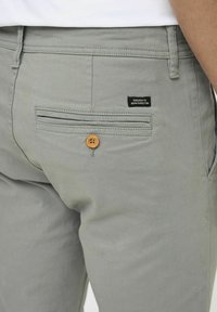 Lichtgrijze broek van een katoenmix, met een achterzak met een houten knoop en een klein zwart label met tekst.